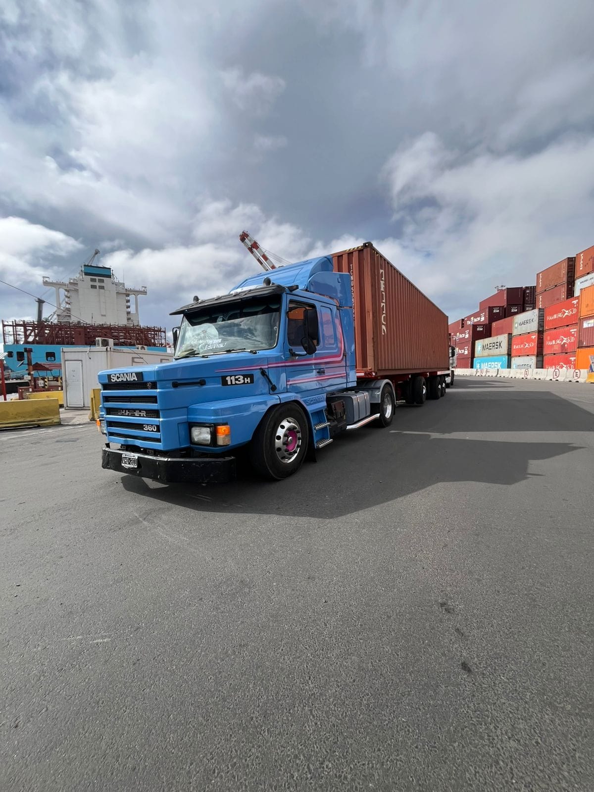 Camión Scania 113H azul transportando un contenedor en el puerto, junto a una grúa